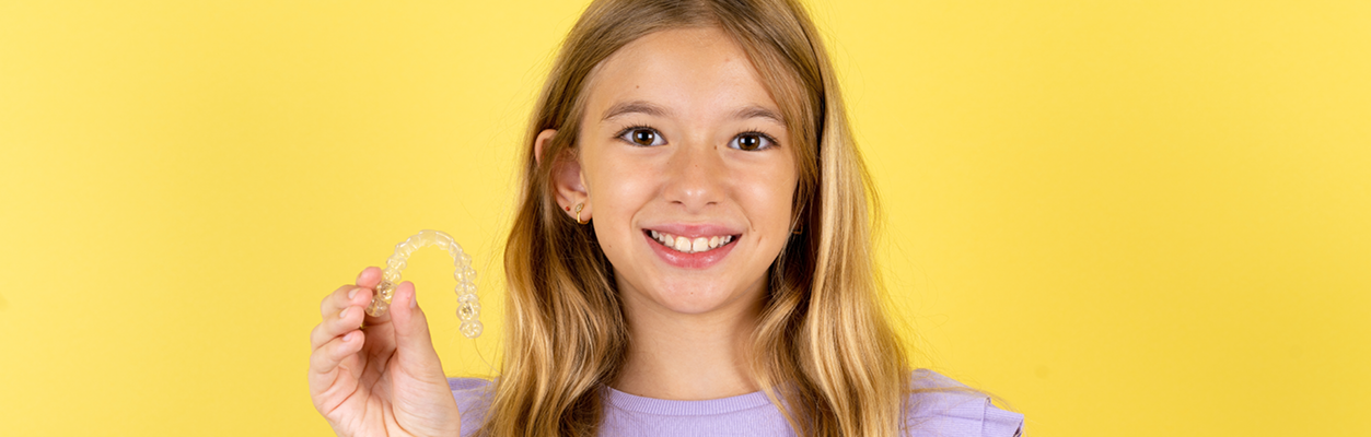 Close up of a young girl with long blonde hair and a light purple shirt, smiling and holding an Invisalign tray.
